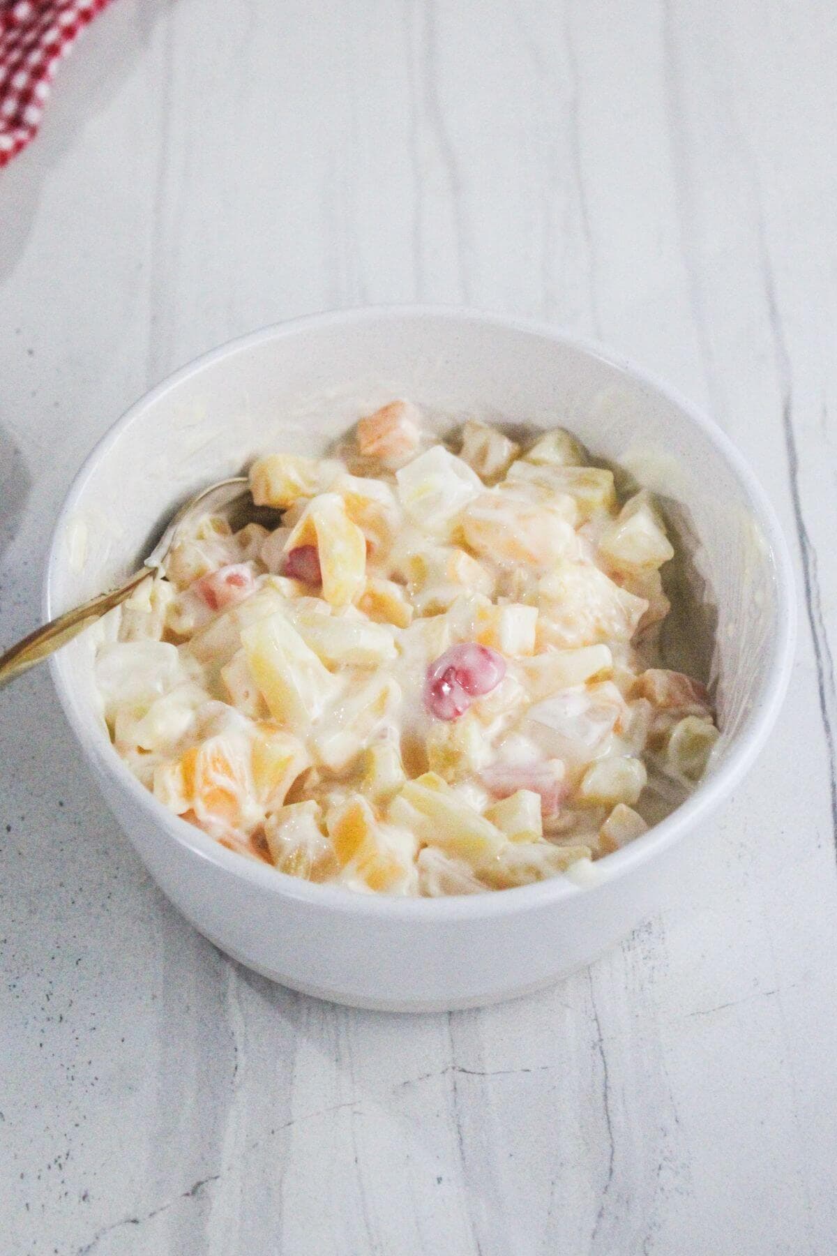 A white bowl filled with creamy fruit salad made of diced fruits and a spoon resting inside, placed on a light-colored surface.