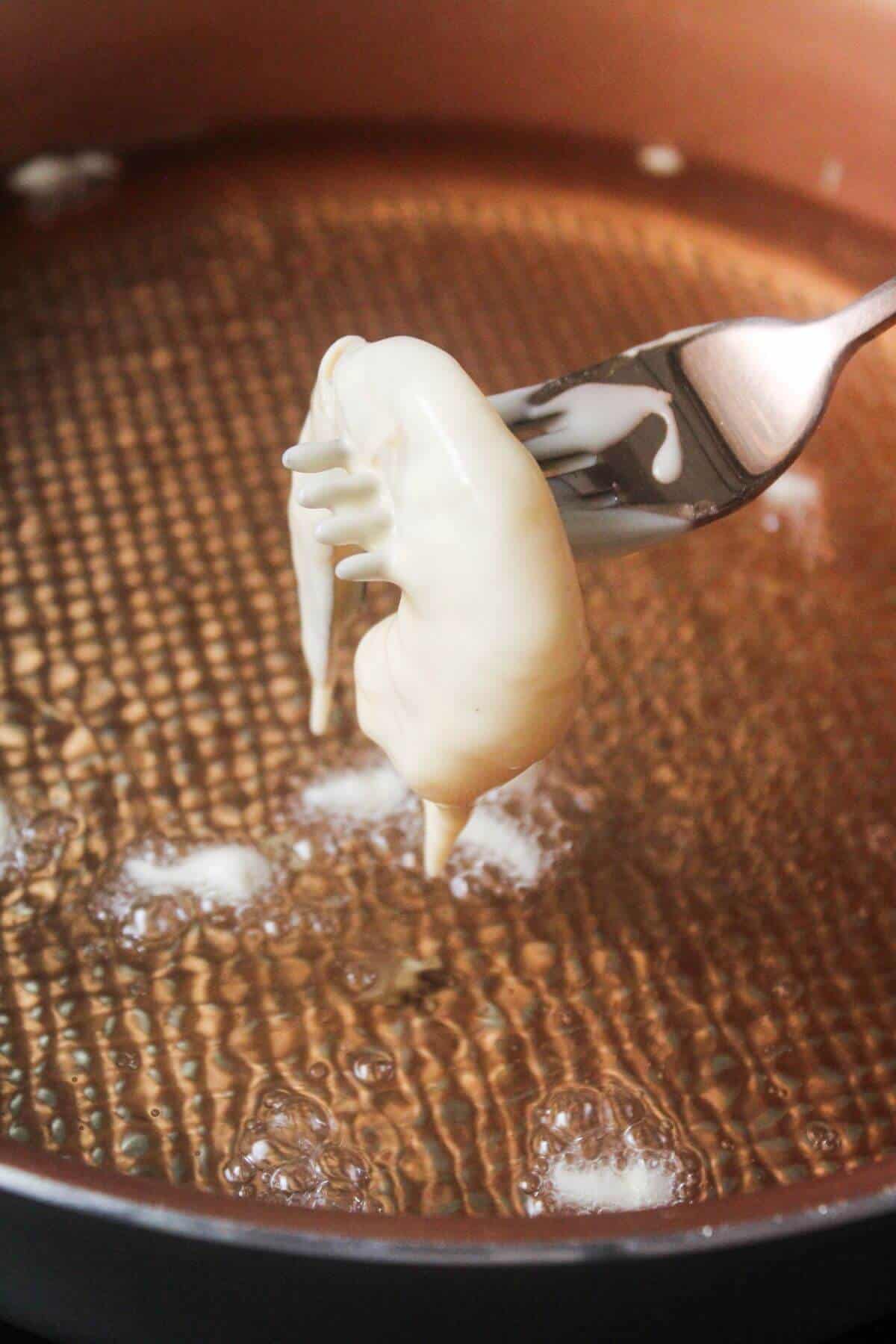 A fork holds a white batter-coated shrimp above a copper-colored pan with oil and bubbles.