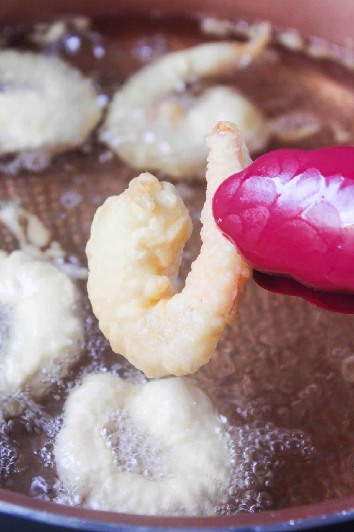 A person wearing a pink glove holds a battered shrimp above hot oil, with other pieces frying in the pan.