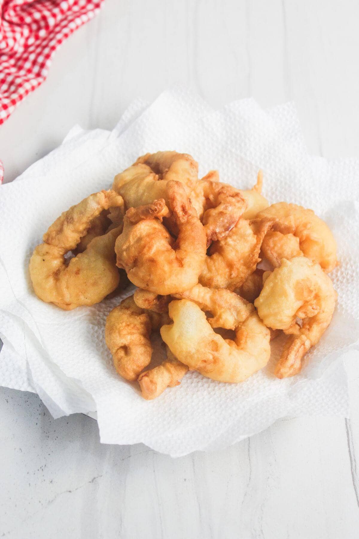 A pile of golden-brown fried shrimp sits on a white paper towel, with a red and white checked cloth in the background.