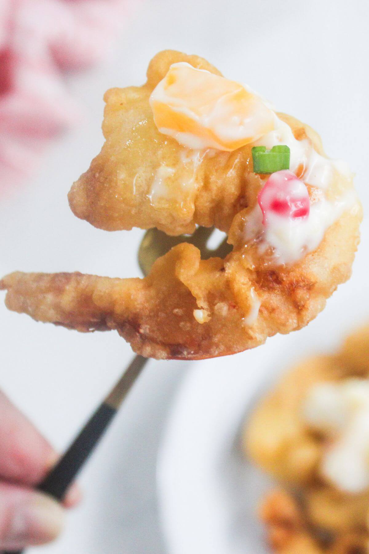A close-up of a fried shrimp topped with creamy sauce, diced red pepper, a slice of cheese, and a piece of green onion, held by a fork.