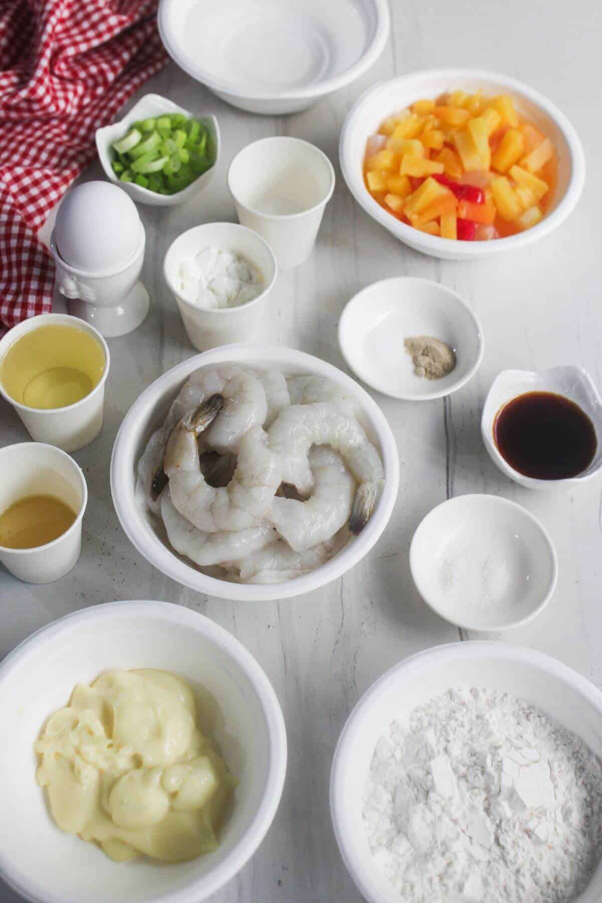 Various ingredients in bowls on a white surface, including raw shrimp, flour, mayonnaise, chopped vegetables, seasonings, and an egg, with a red checkered cloth partially visible.