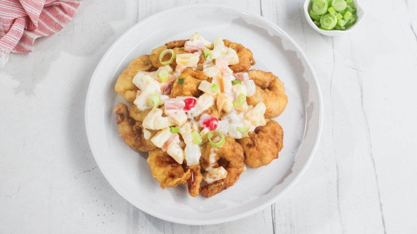 A white plate with battered fried shrimp topped with a creamy salad of chopped vegetables and sliced green onions, on a white surface.