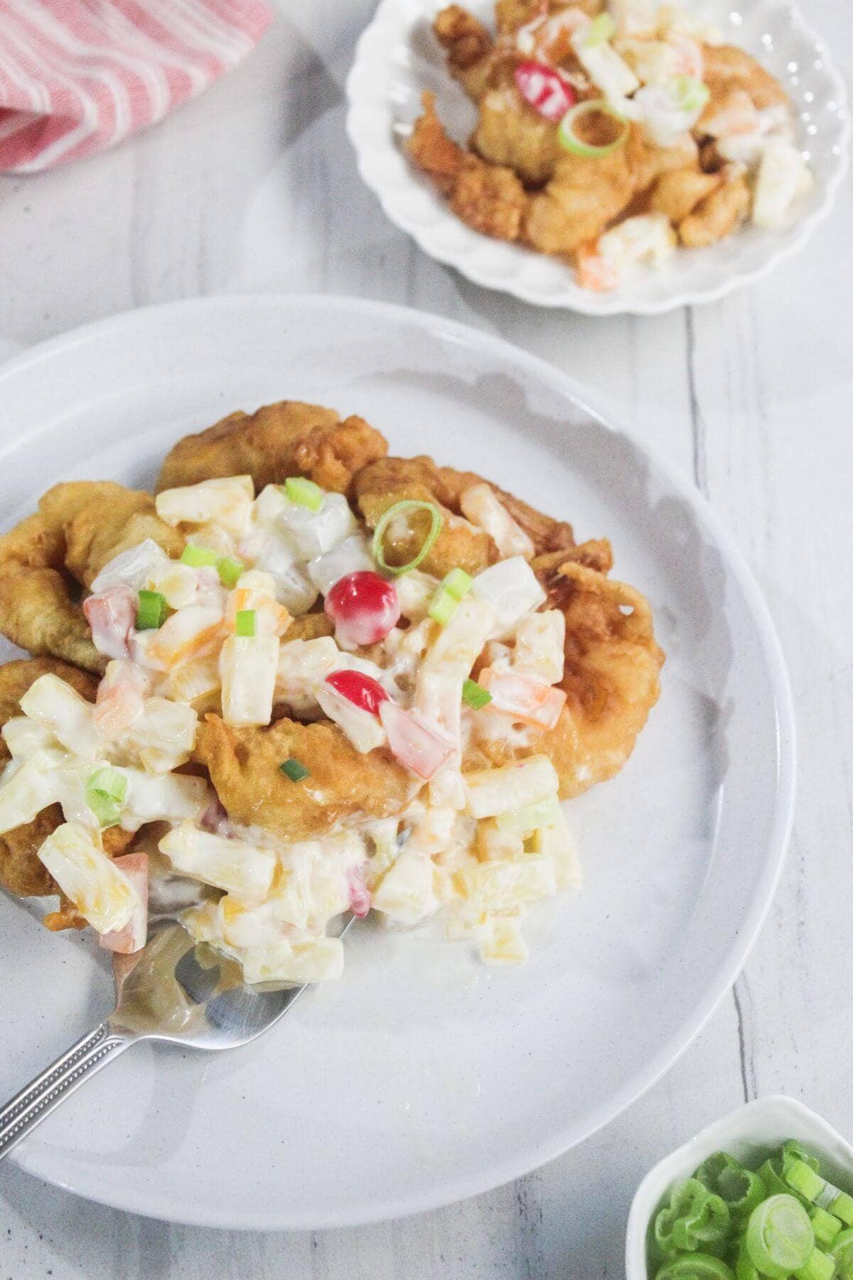 A plate of fried chicken fillets topped with a creamy fruit and vegetable salad, garnished with chopped green onions and a cherry.