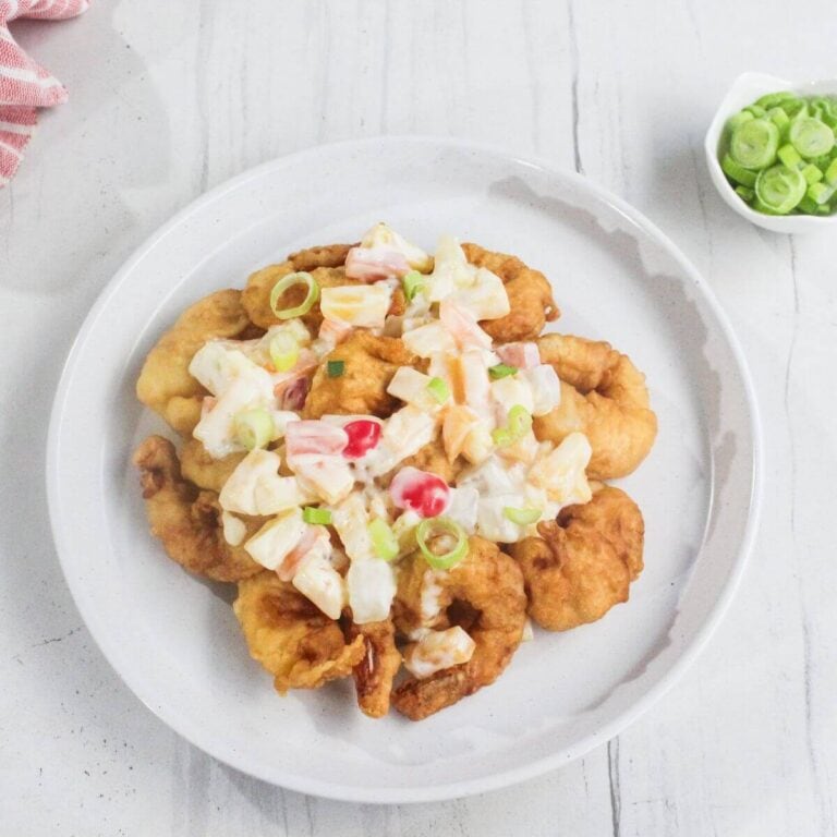 A white plate with pieces of fried chicken topped with a creamy salad of diced vegetables and garnished with sliced green onions.