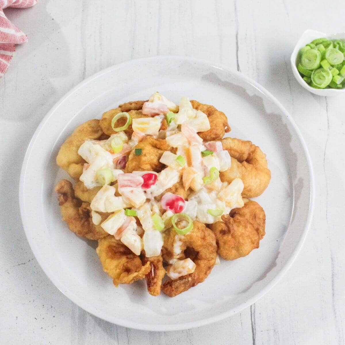 A white plate with pieces of fried chicken topped with a creamy salad of diced vegetables and garnished with sliced green onions.