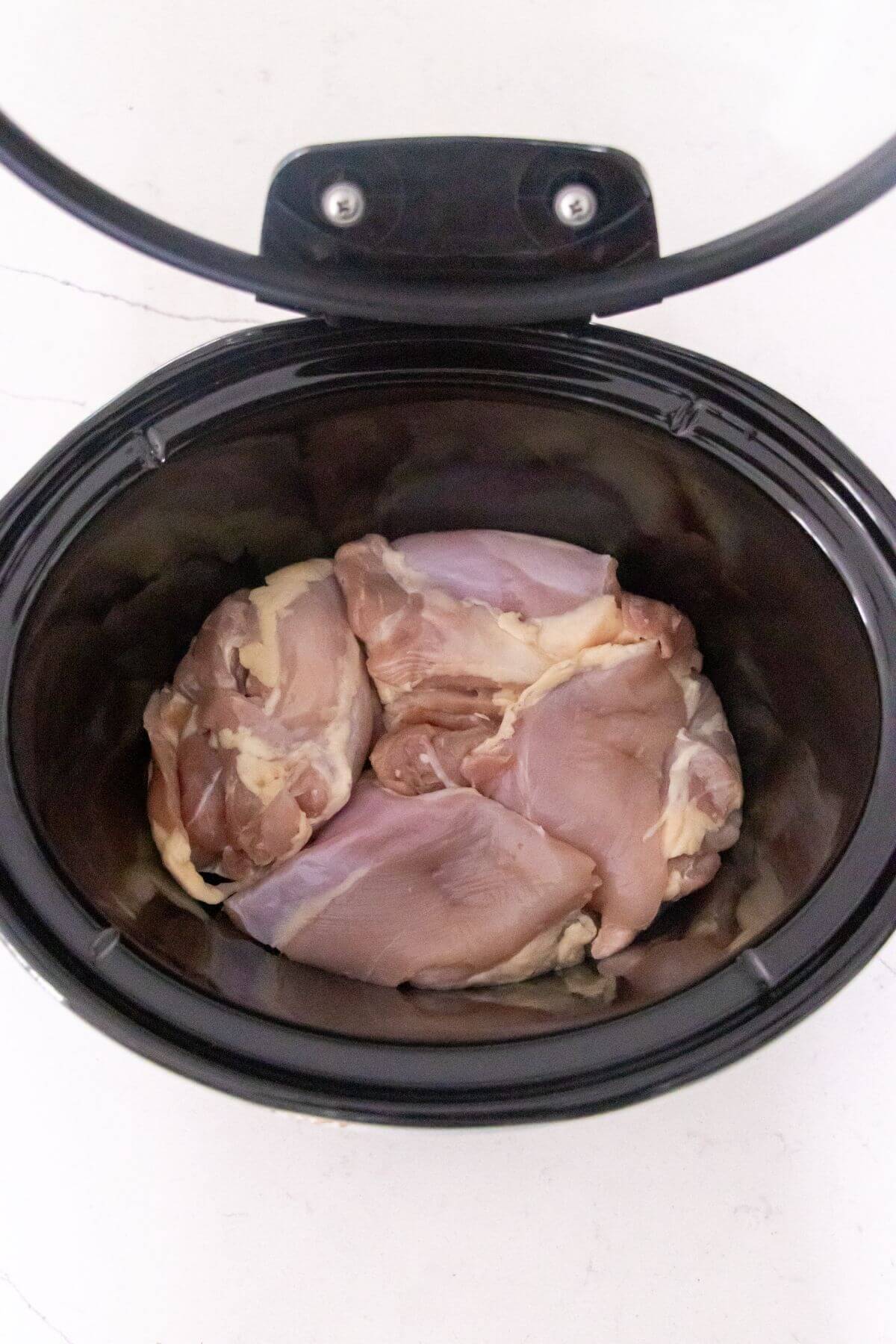 Raw chicken thighs placed inside a black slow cooker on a white surface.
