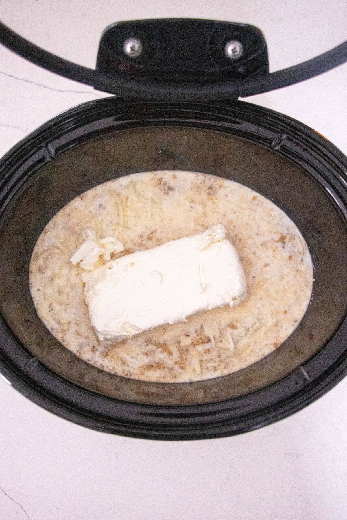 A slow cooker containing a block of cream cheese, shredded cheese, ground meat, and a creamy liquid mixture.
