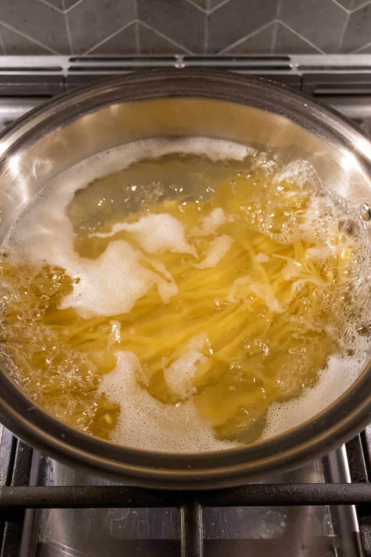 A stainless steel pot of pasta boiling in water on a stovetop with bubbles and foam forming on the surface.