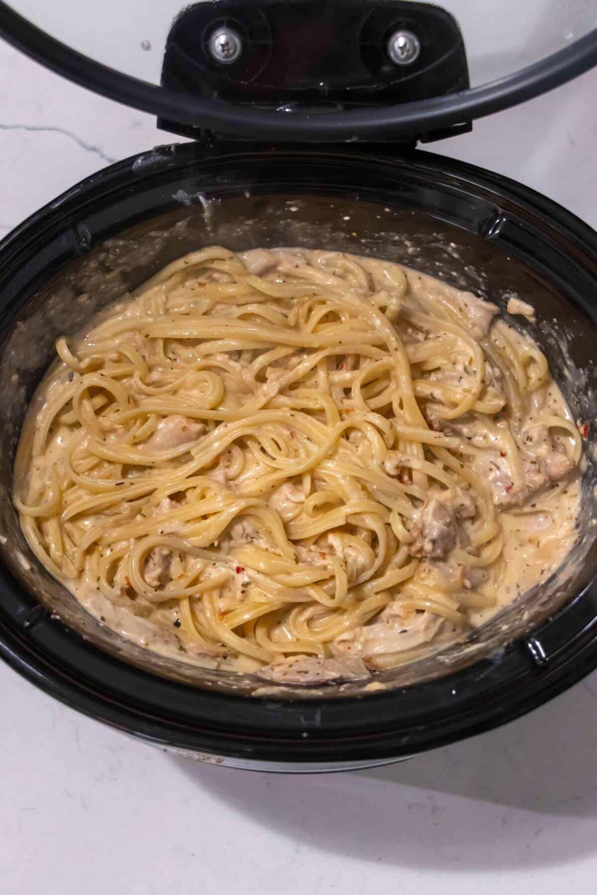 Cooked spaghetti mixed with a creamy sauce and pieces of chicken inside a slow cooker with the lid open.