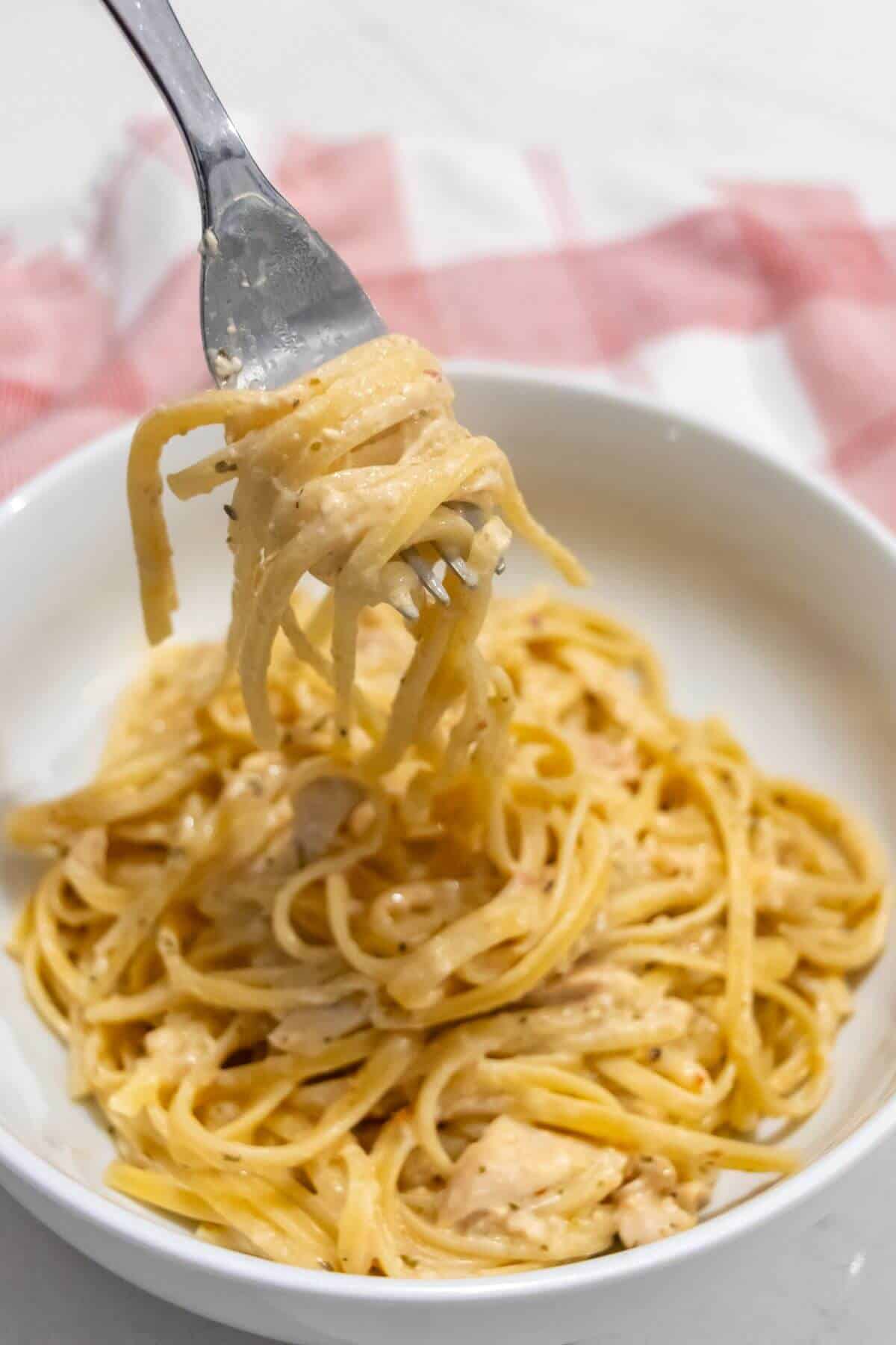 A fork lifts a serving of creamy pasta from a white bowl filled with linguine in a light sauce, with a red and white cloth in the background.
