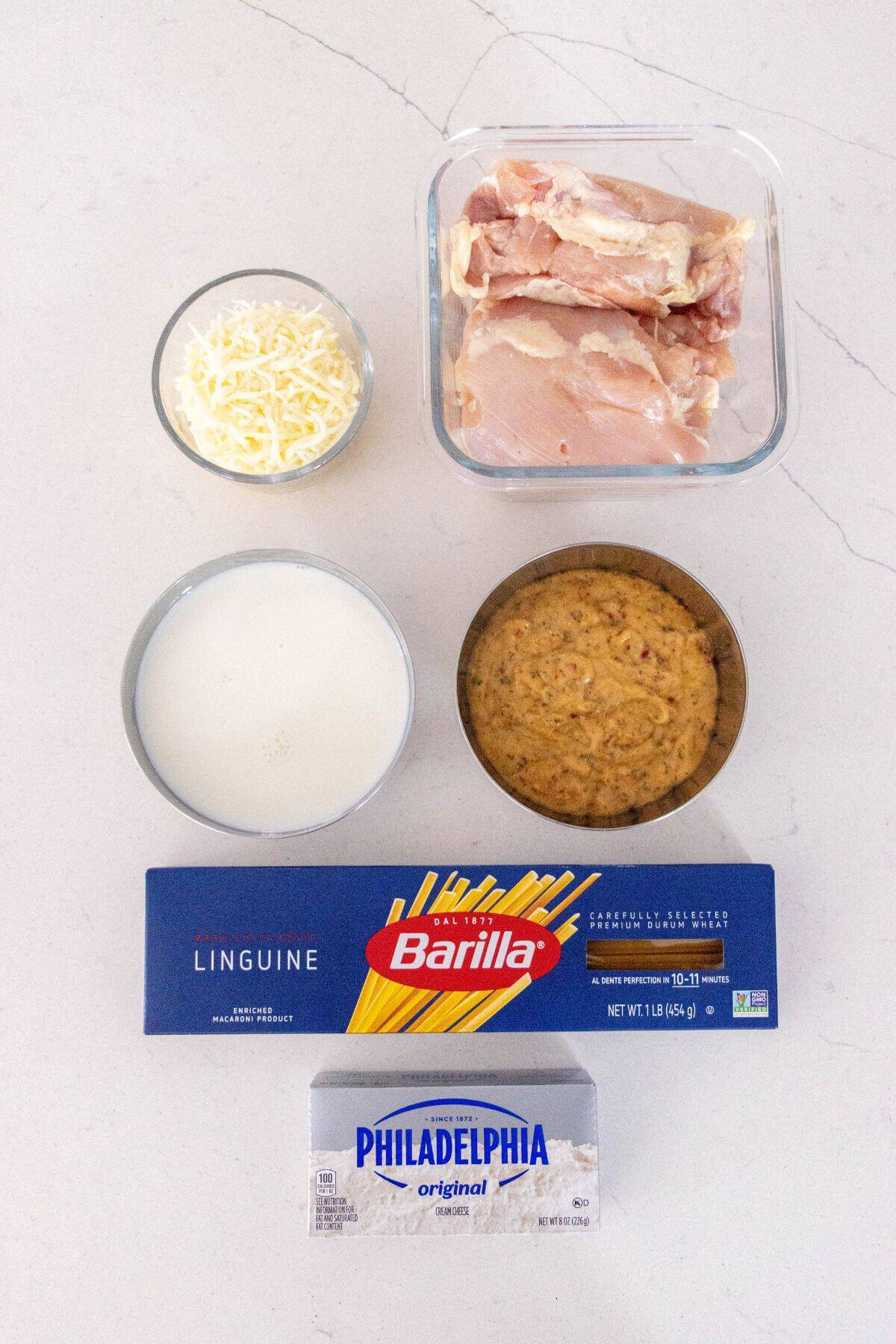 Top-down view of uncooked linguine, chicken thighs, milk, shredded cheese, cream cheese, and a bowl of sauce on a white surface.