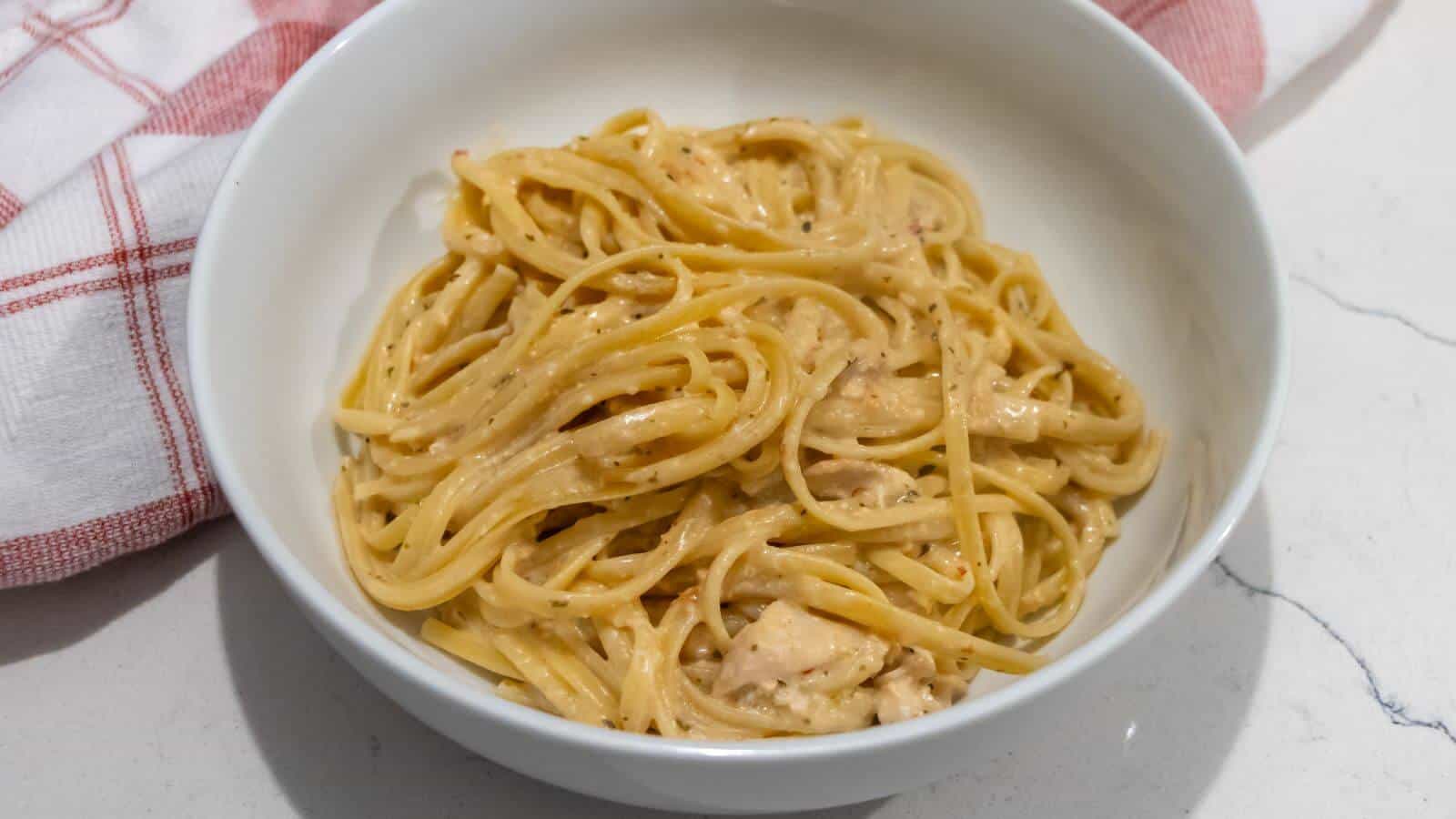 A white bowl filled with creamy pasta, likely spaghetti or linguine, placed on a light countertop next to a red and white checkered cloth.