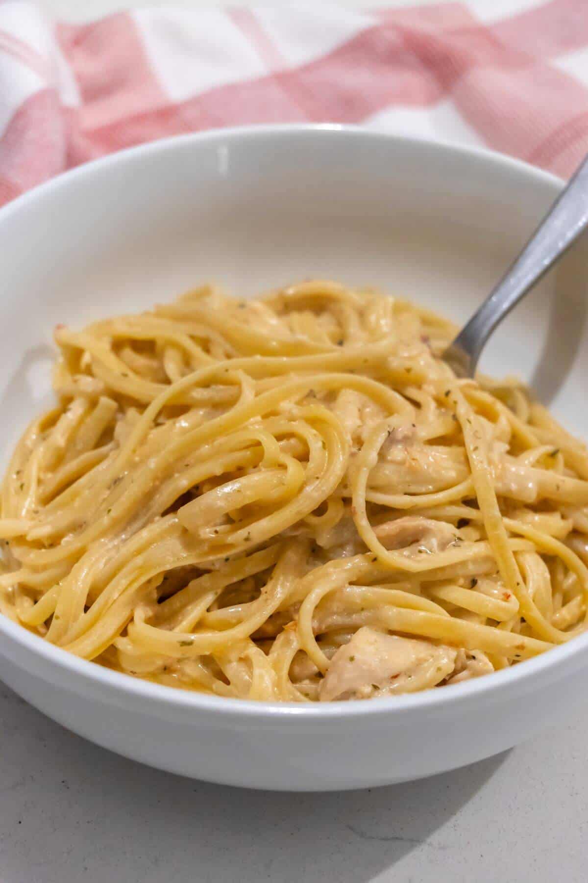 A white bowl filled with creamy spaghetti, with a fork resting in the noodles. A red and white cloth is partially visible in the background.