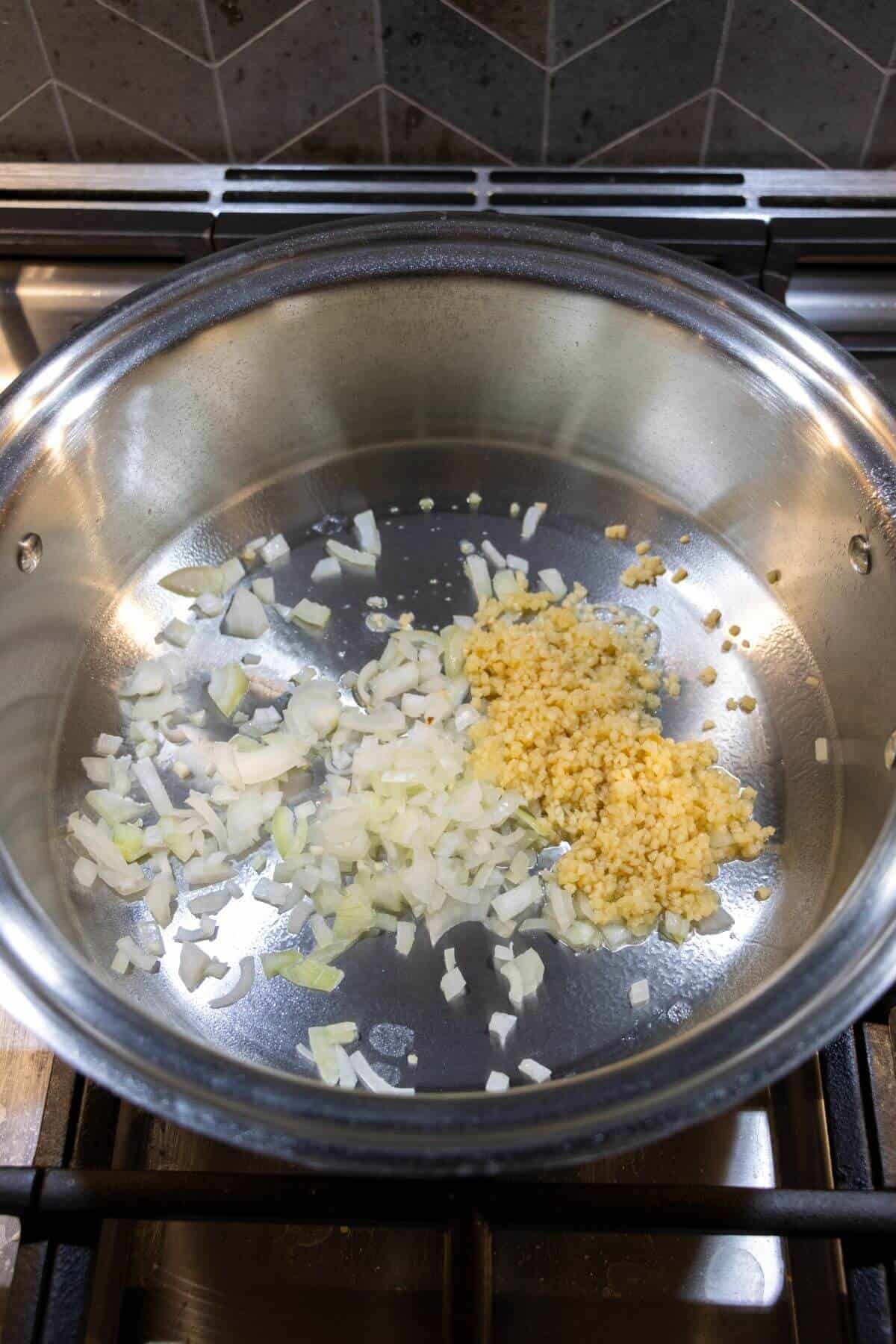 Chopped onions and minced garlic are being sautéed in a stainless steel pan on a stovetop.