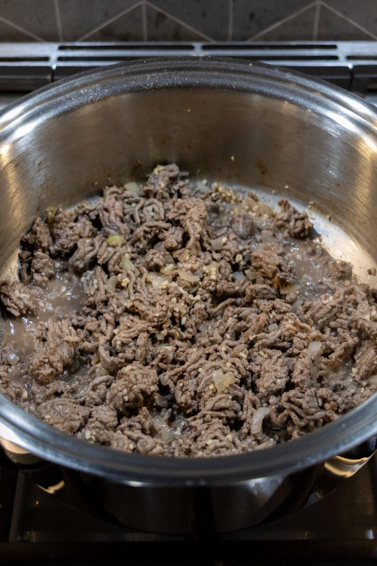 Ground beef being browned with chopped onions and garlic in a stainless steel pan on a stovetop.