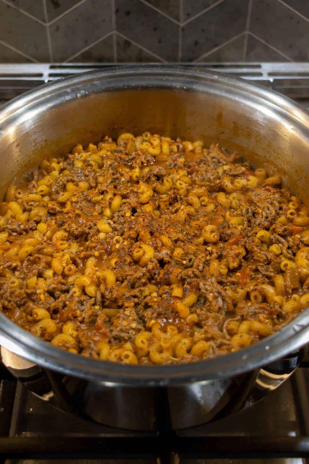Large pot of cooked macaroni mixed with ground beef and tomato sauce sits on a stovetop, with a tiled backsplash in the background.