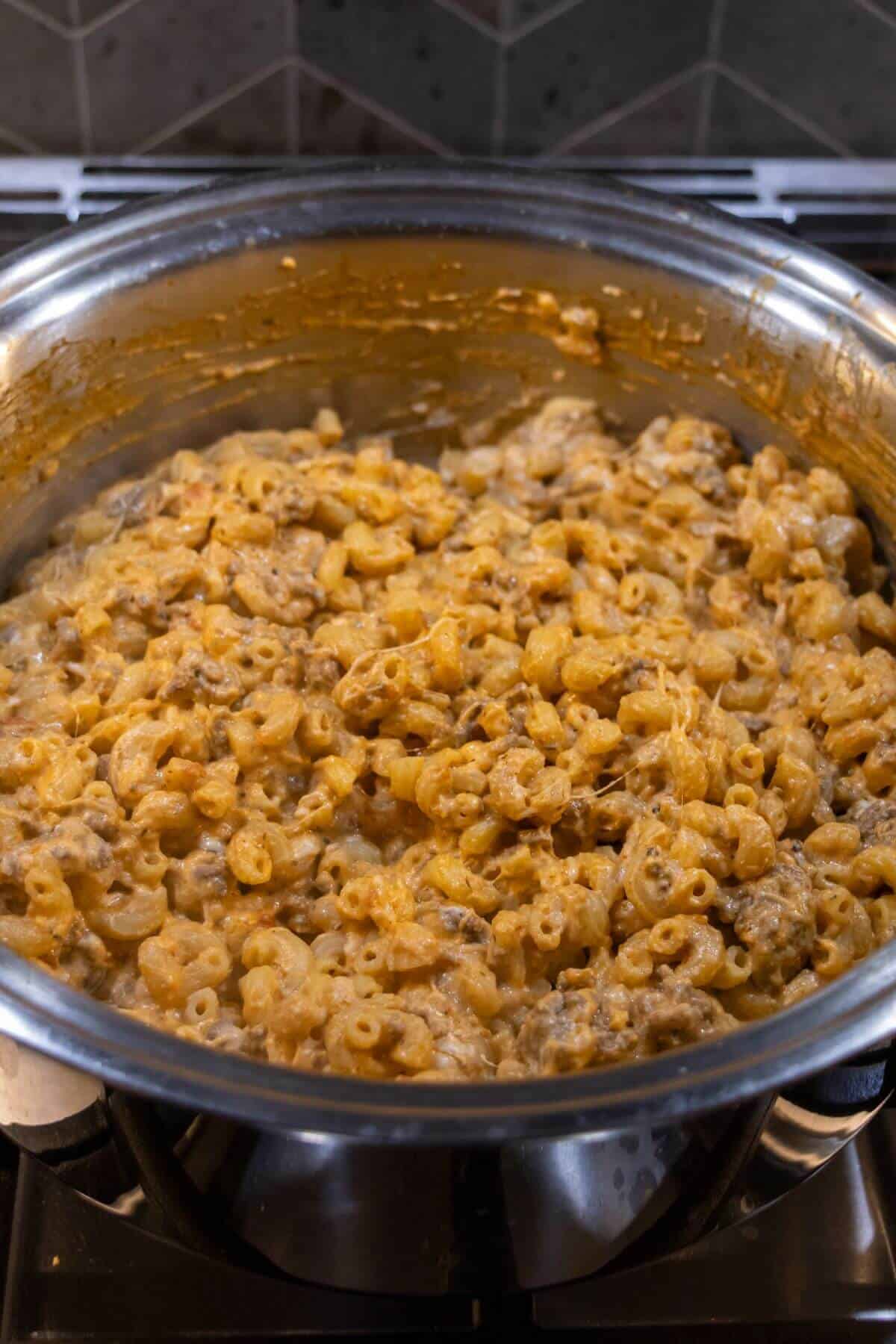 A pot filled with cooked macaroni mixed with ground meat and a creamy, orange-colored sauce sits on a stovetop.