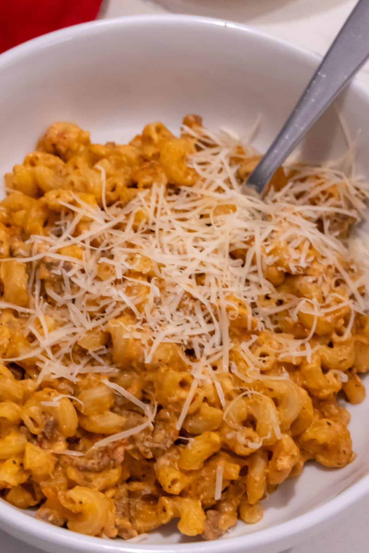 A white bowl filled with macaroni and cheese topped with shredded cheese, with a metal spoon placed inside.