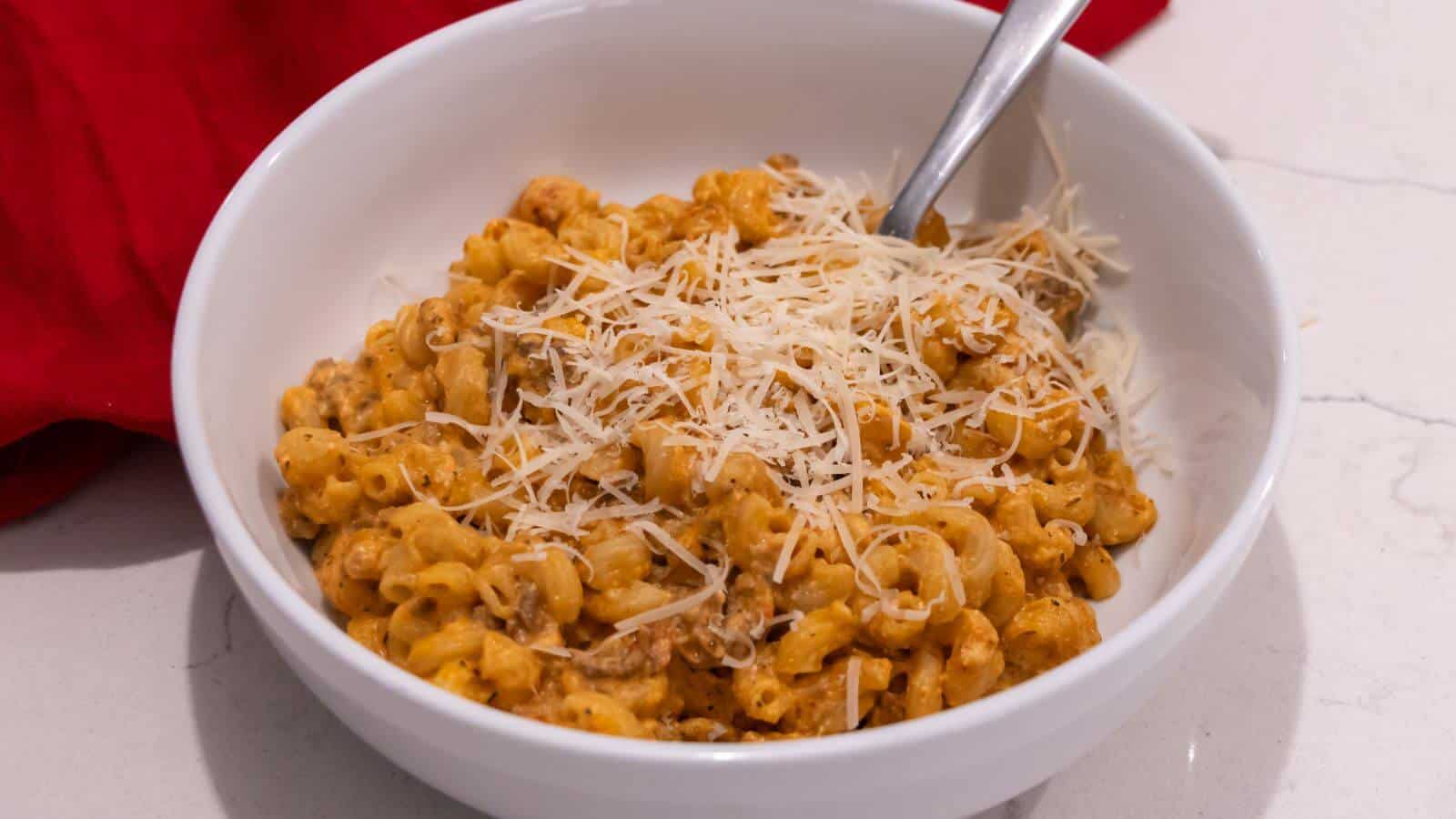 A white bowl filled with macaroni and ground beef in a creamy sauce, topped with shredded cheese, sits on a white countertop with a spoon and a red cloth nearby.