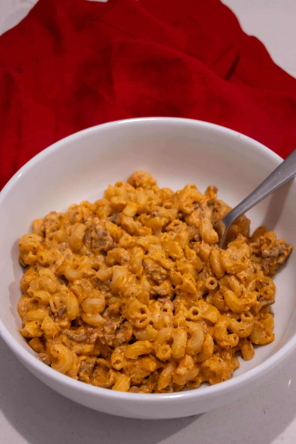 A white bowl filled with macaroni and cheese mixed with ground beef, with a metal spoon inside. A red cloth is in the background.