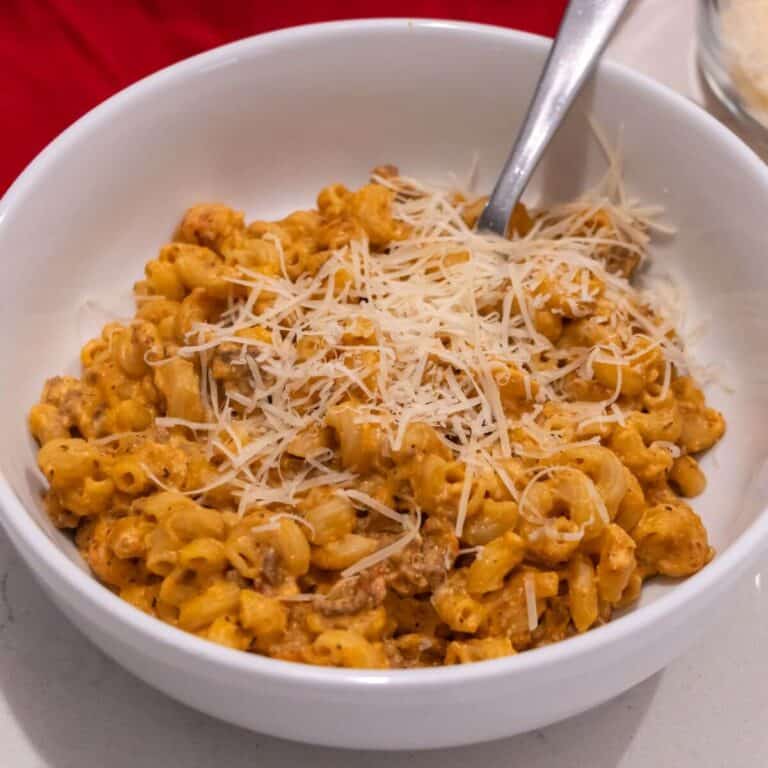 A white bowl filled with macaroni mixed with a creamy orange sauce and ground meat, topped with shredded cheese, with a spoon in the bowl.