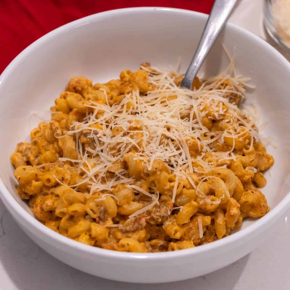A white bowl filled with macaroni mixed with a creamy orange sauce and ground meat, topped with shredded cheese, with a spoon in the bowl.