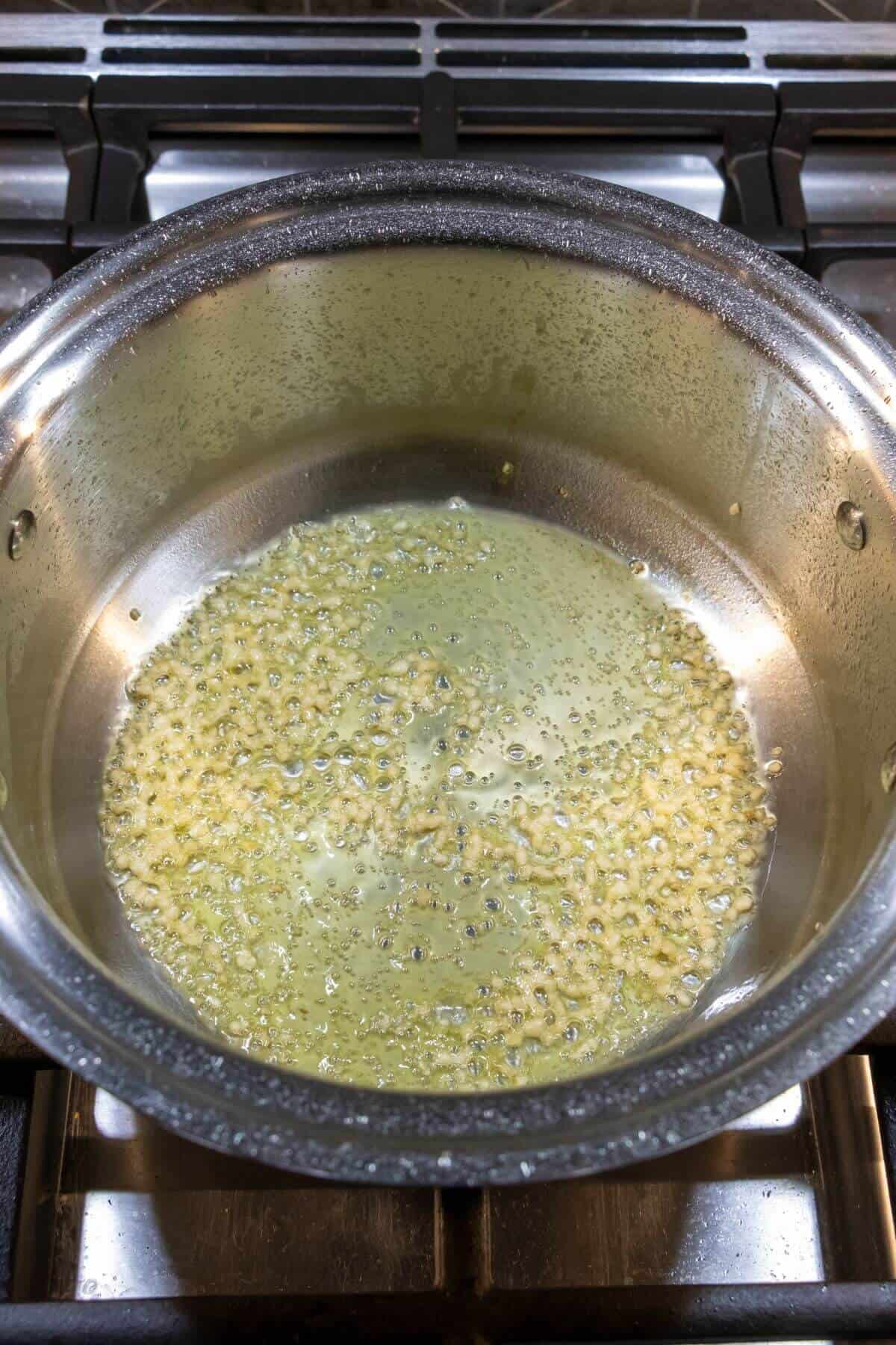 Chopped garlic sautéing in oil in a stainless steel pot on a stovetop.