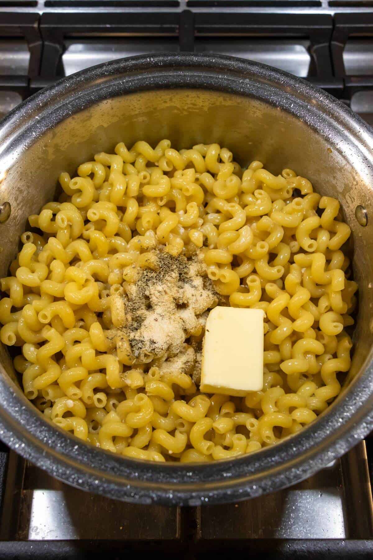 Cooked macaroni pasta in a pot with a pat of butter and seasonings on top, sitting on a stovetop.