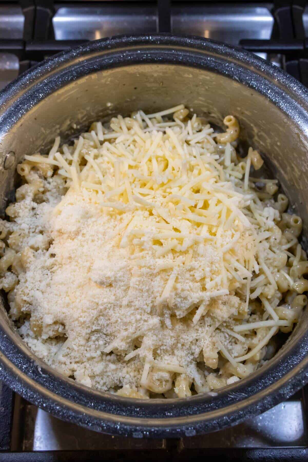 A pot of cooked macaroni on a stovetop topped with shredded cheese and grated Parmesan, ready to be mixed.