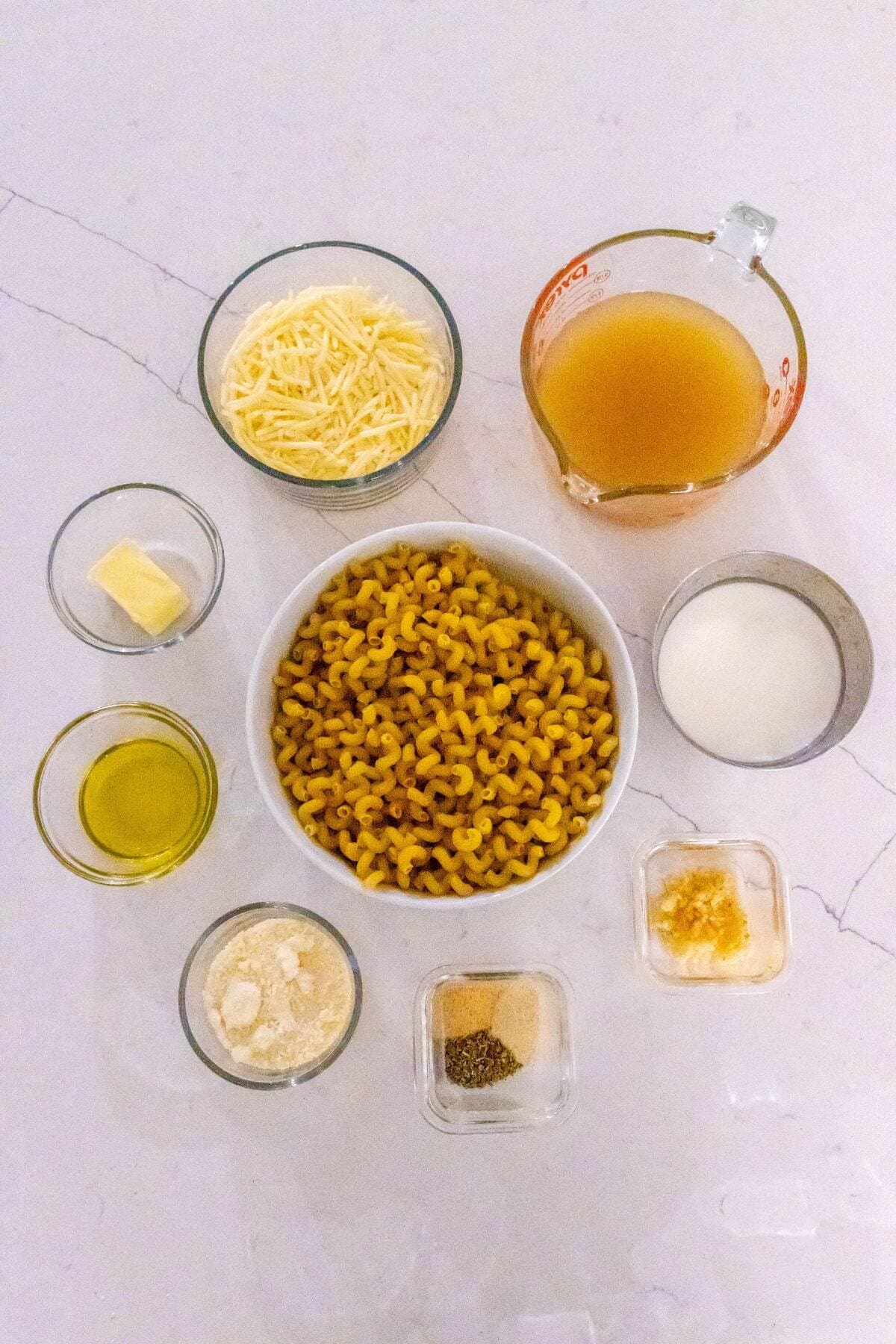 Overhead view of measured ingredients for macaroni and cheese, including pasta, grated cheese, broth, milk, butter, olive oil, spices, and seasonings on a white countertop.