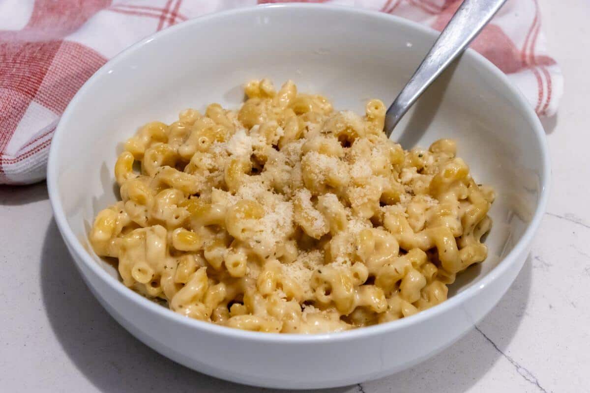 A white bowl of macaroni and cheese topped with grated parmesan, with a spoon in the bowl and a red and white cloth in the background.