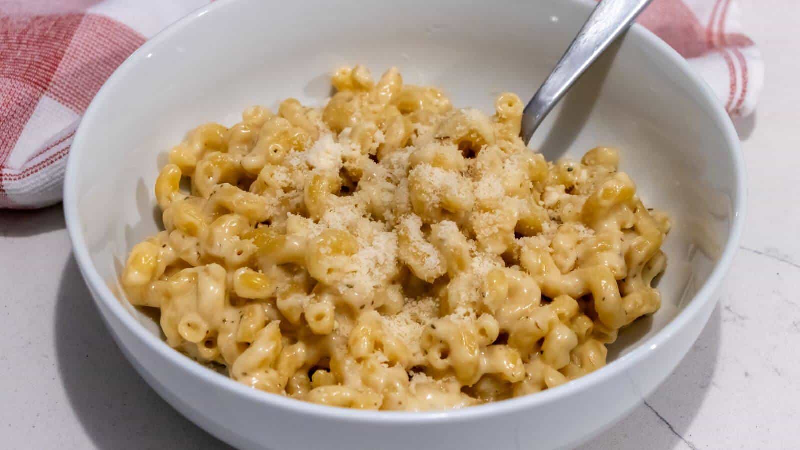A bowl of macaroni and cheese topped with grated parmesan, with a spoon placed inside the bowl. A red and white cloth is partially visible in the background.