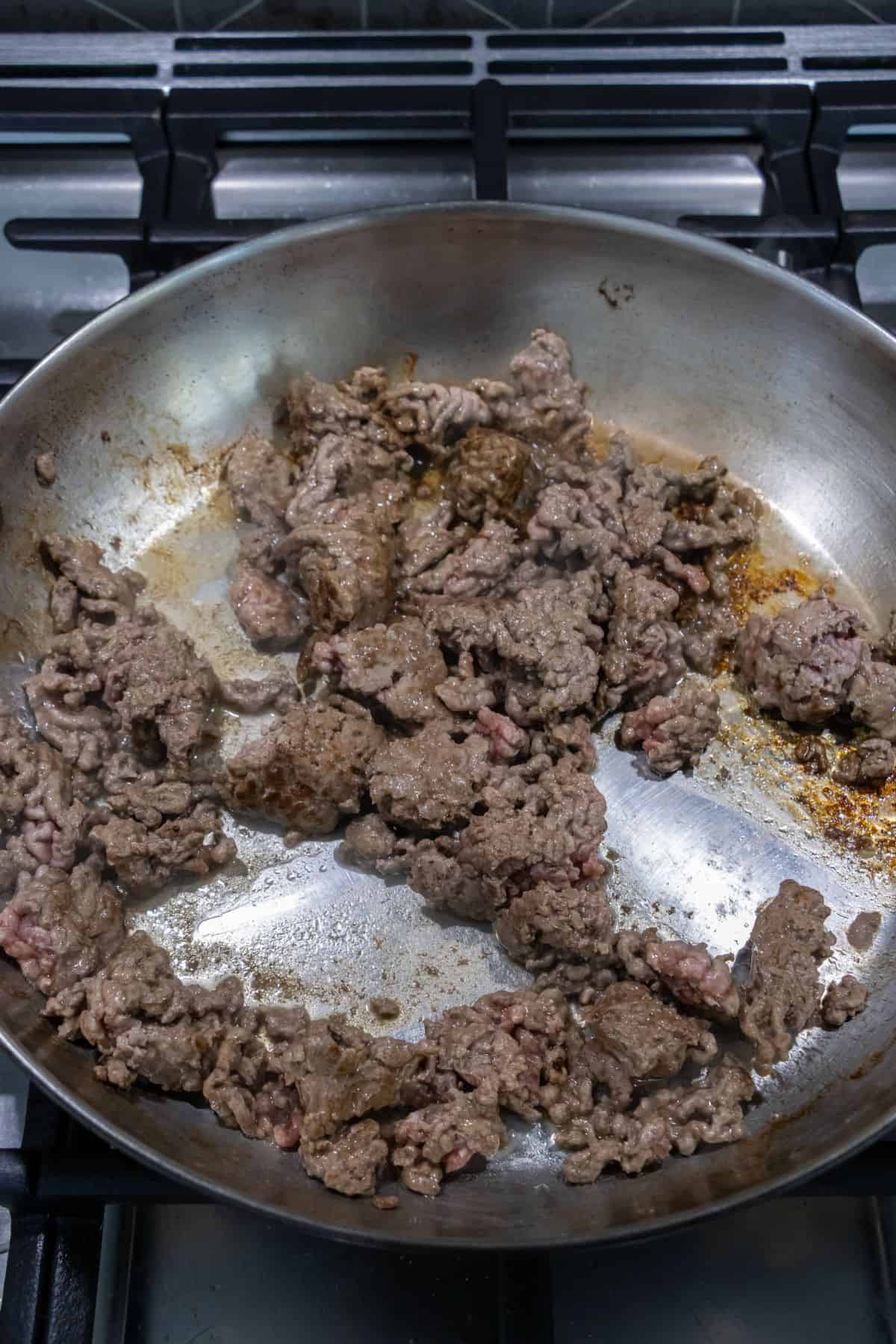 Ground beef cooking in a stainless steel skillet on a stovetop, with some pieces browned and others still pink.