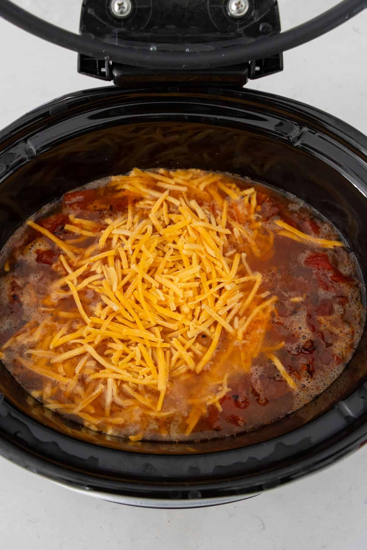 A slow cooker filled with soup ingredients, including shredded cheddar cheese on top, before cooking.