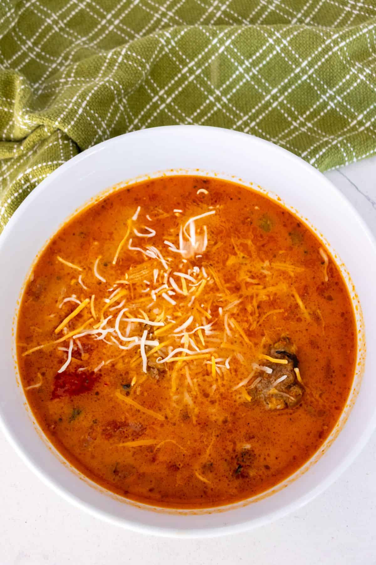 A white bowl filled with creamy orange soup topped with shredded cheese, placed on a white surface beside a green checkered cloth.