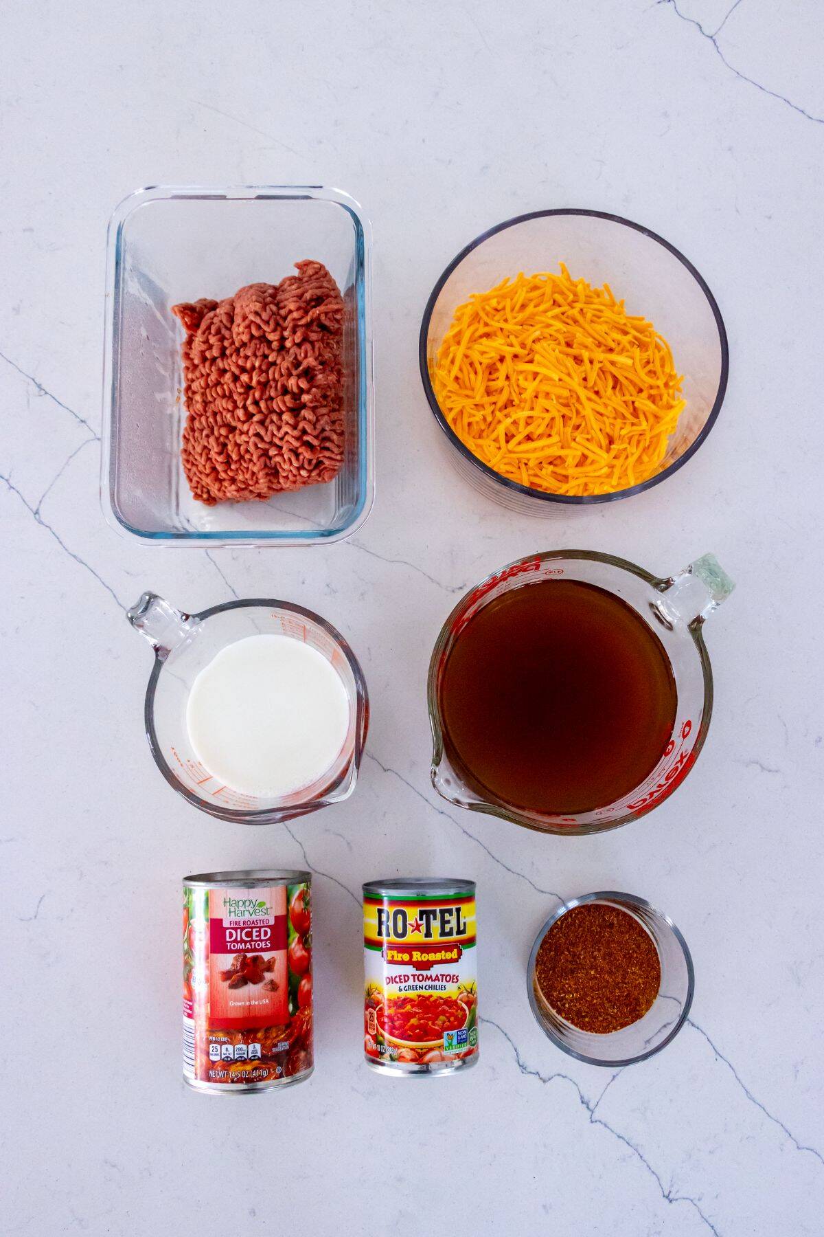 Ground beef, shredded cheddar cheese, milk, beef broth, taco seasoning, a can of diced tomatoes, and a can of diced tomatoes with green chilies are arranged on a white surface.