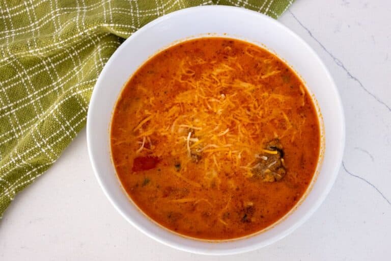 A white bowl filled with orange-colored soup topped with shredded cheese, placed on a white surface next to a green checkered cloth.