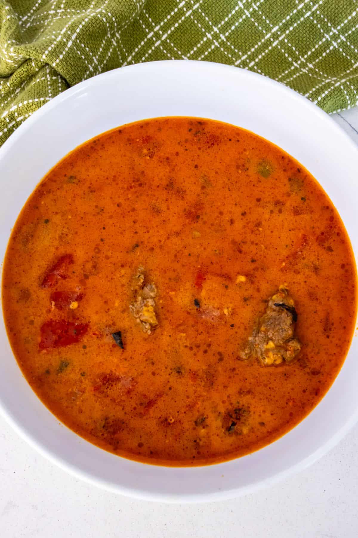 A bowl of reddish soup with visible chunks of meat and tomatoes, set on a white surface next to a green and white checkered cloth.