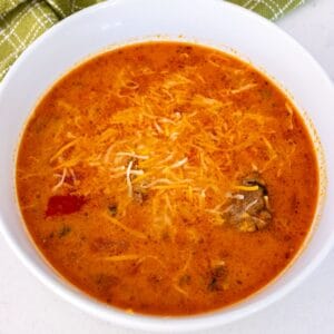 A bowl of creamy orange soup topped with shredded cheese, containing visible chunks of vegetables and meat, on a white surface with a green cloth in the background.
