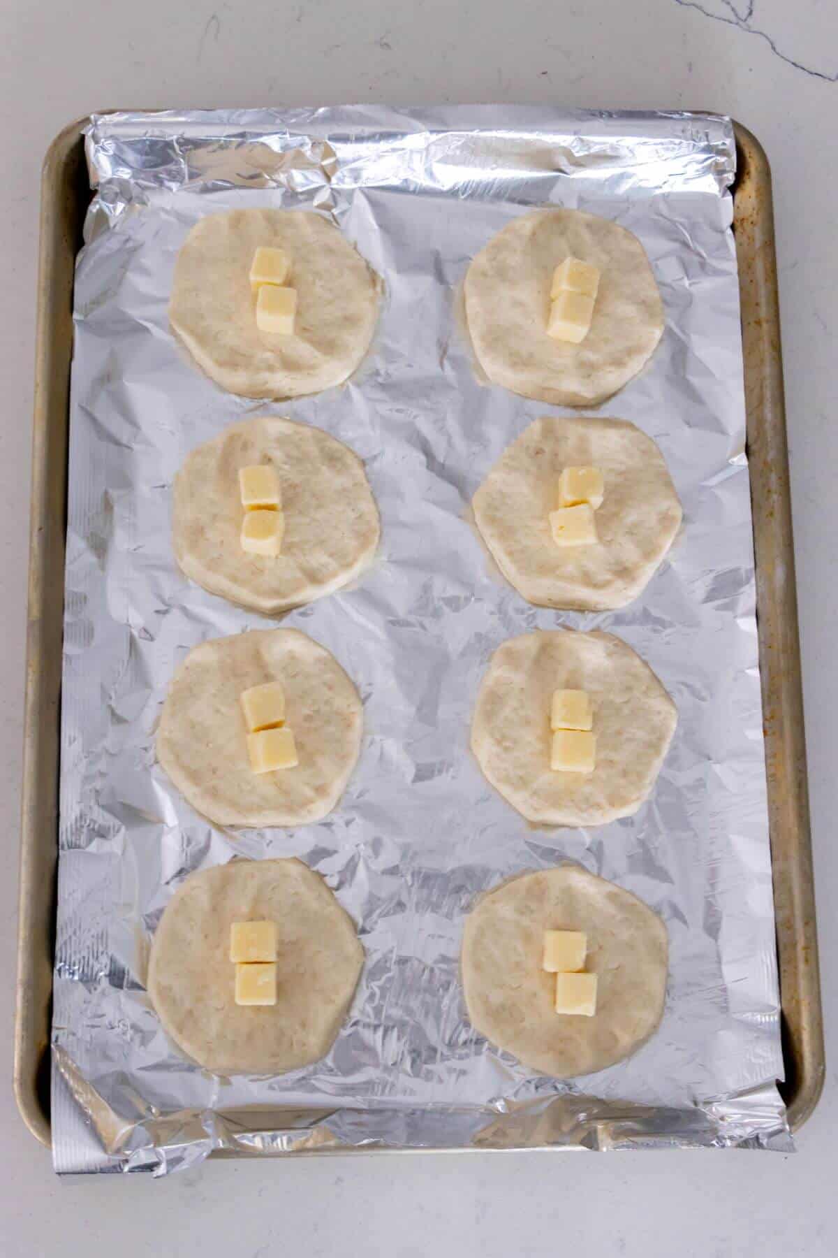 A baking sheet lined with foil holds eight rounds of uncooked dough, each topped with two small cubes of butter.