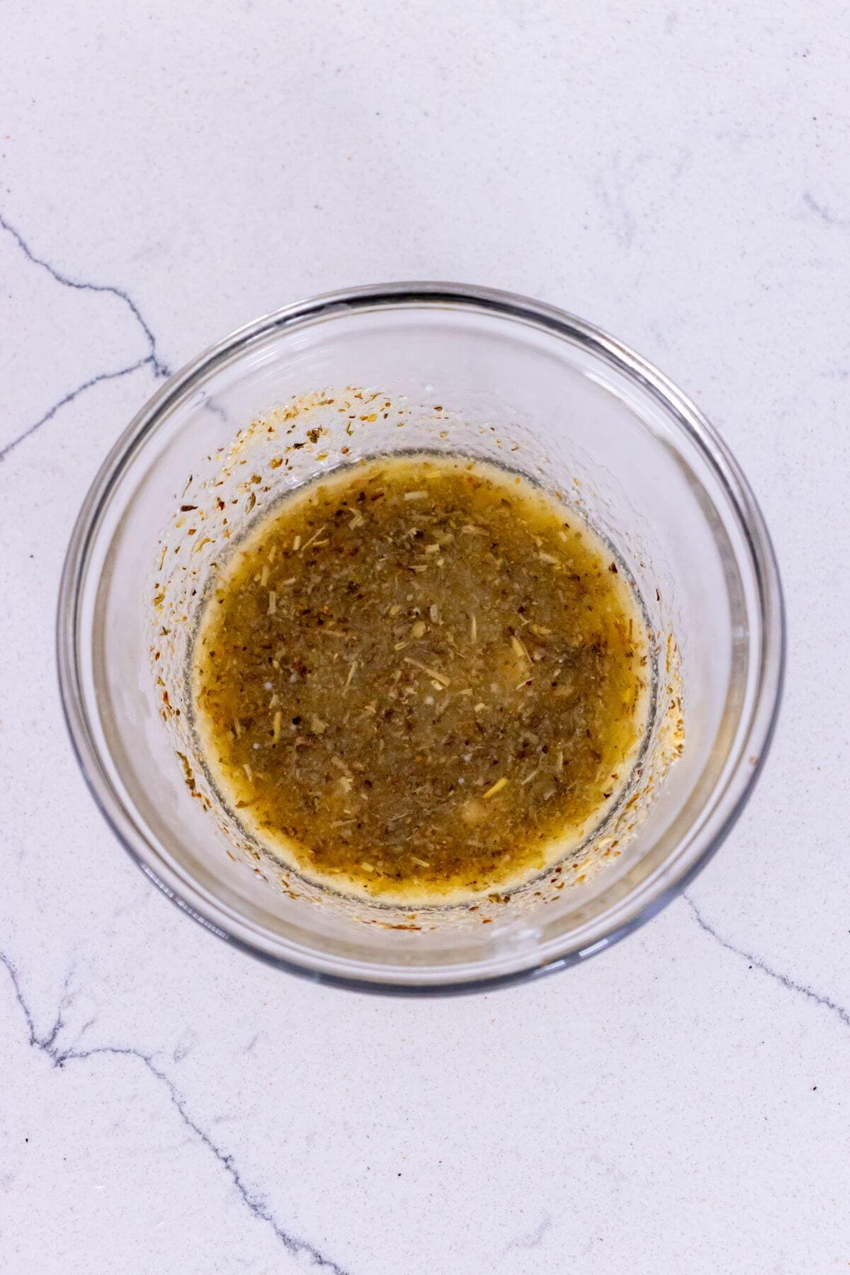 A clear glass bowl filled with a yellowish, seasoned liquid mixture sits on a white marble surface.