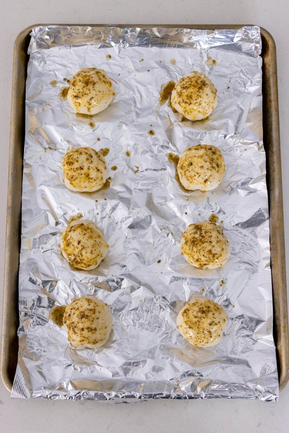 Eight dough balls sprinkled with seasoning are arranged on a foil-lined baking sheet, ready to be baked.