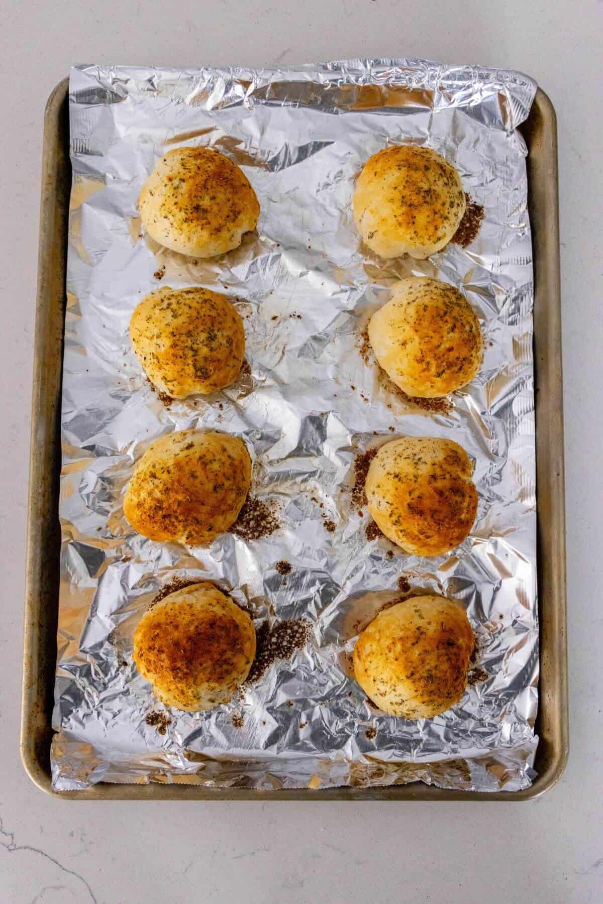 A baking sheet lined with aluminum foil holds eight round, golden-brown rolls sprinkled with herbs and spices.