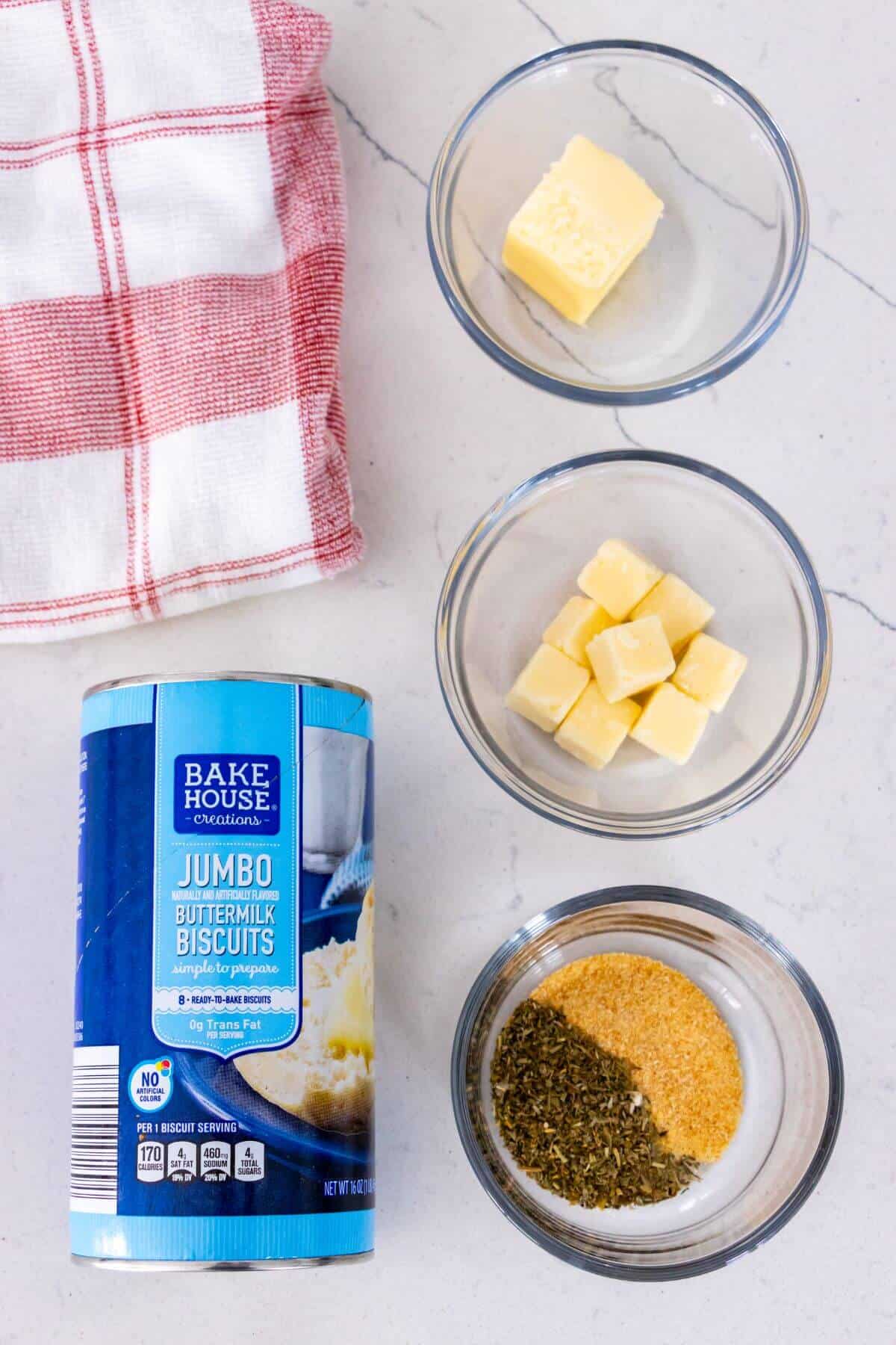 A can of Bake House buttermilk biscuits, cubes of cheese, a pat of butter, a bowl of mixed herbs and seasoning, and a red checked towel on a white surface.