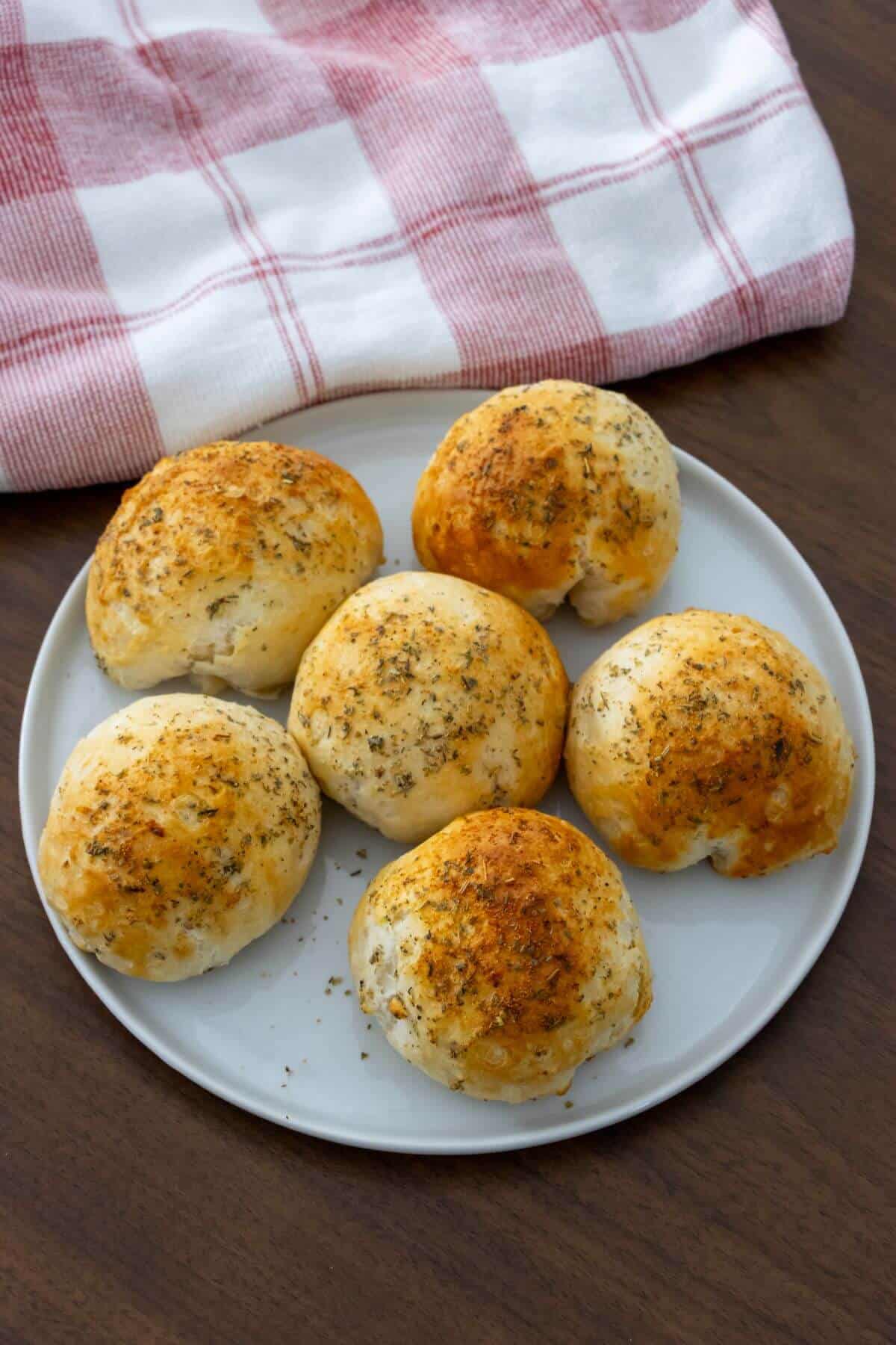 Six golden-brown, seasoned bread rolls on a white plate, with a red and white checkered cloth in the background.