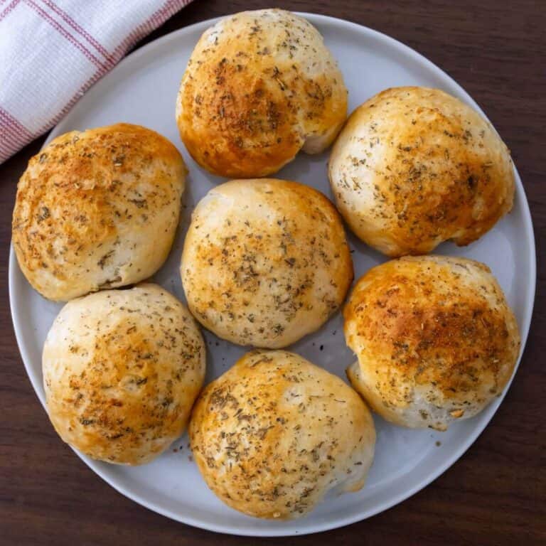 A white plate with seven round, golden-brown bread rolls topped with herbs, placed on a dark wooden surface next to a folded kitchen towel.