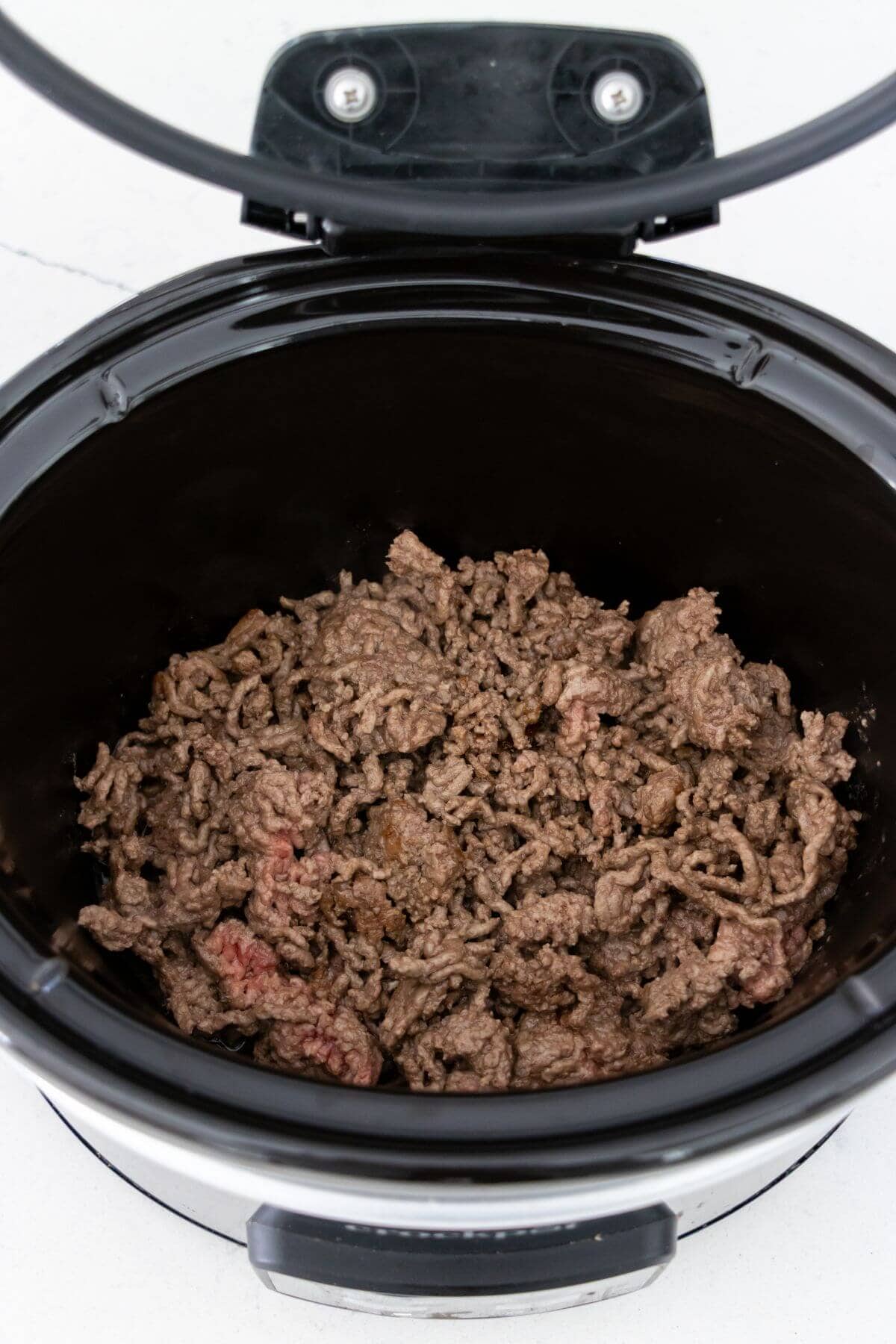 Cooked ground beef inside a black slow cooker with the lid open, showing fully browned meat with some slightly pink areas.