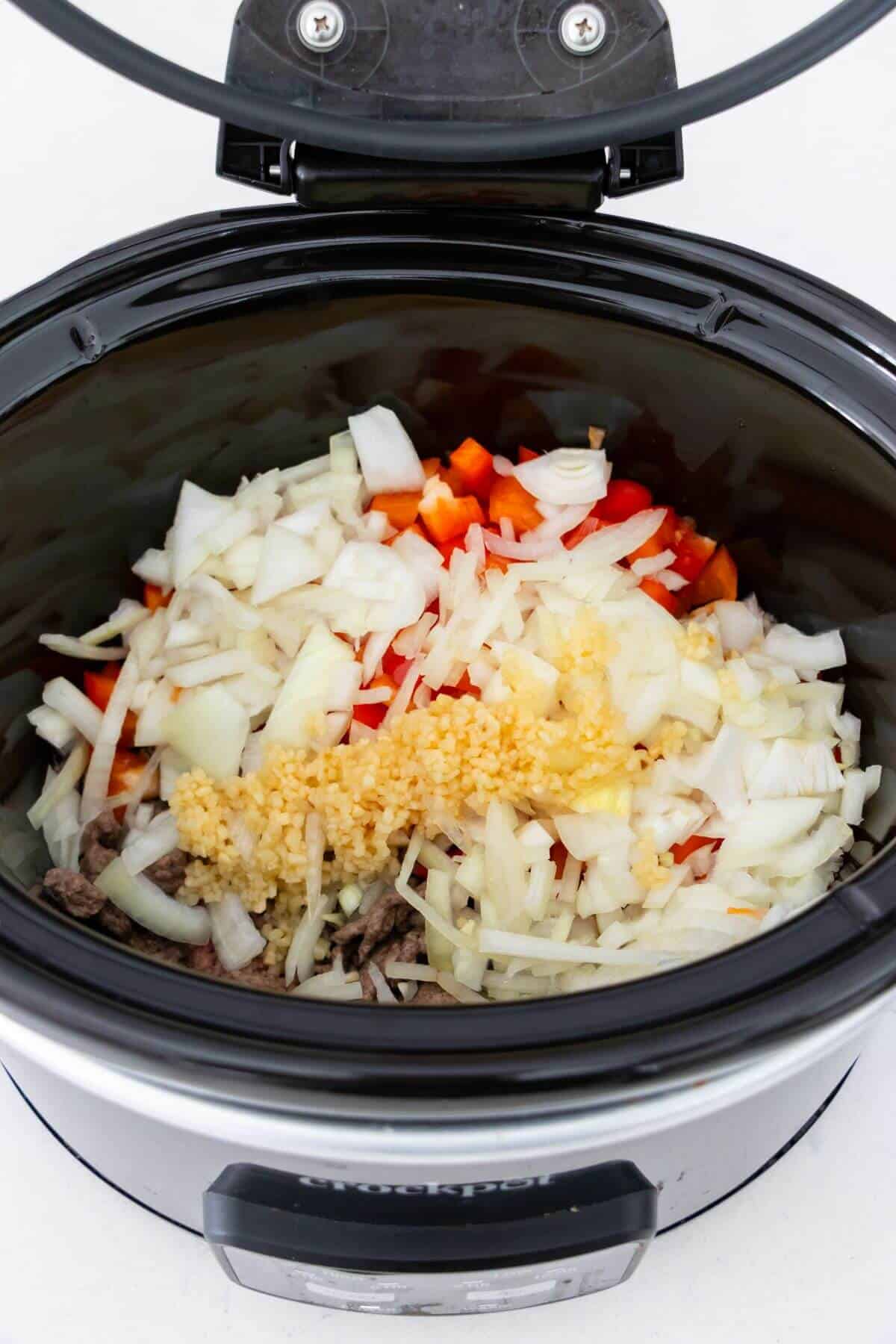 A slow cooker filled with chopped onions, red bell peppers, and minced garlic on top of other ingredients, ready to be cooked.