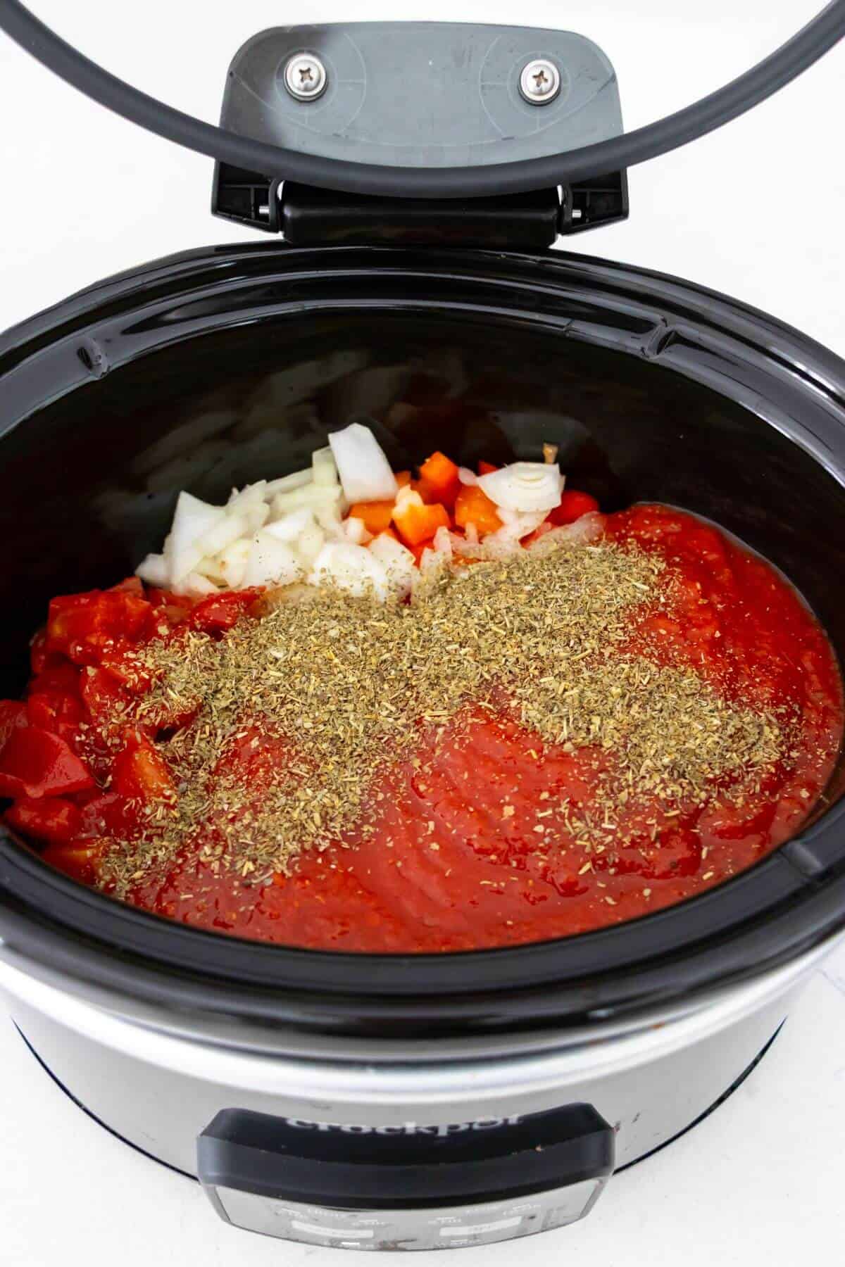 A slow cooker filled with chopped onions, diced red bell peppers, tomato sauce, diced tomatoes, and dried herbs, ready to be cooked.